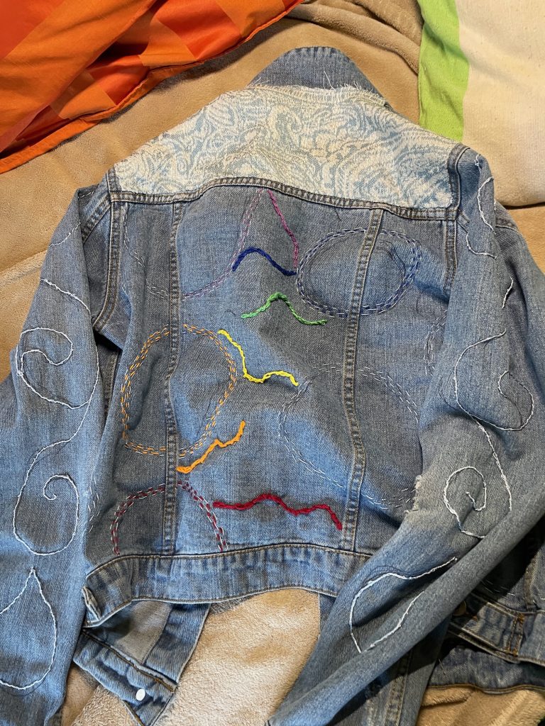 Denim jacket back with colorful hand stitching and white patterned shoulders.